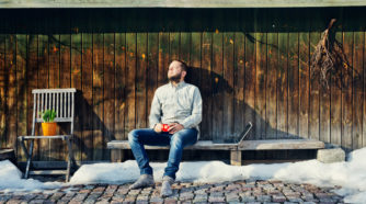 Mies istuu talon seinustalla kahvikuppi kädessä ja ottaa aurinkoa