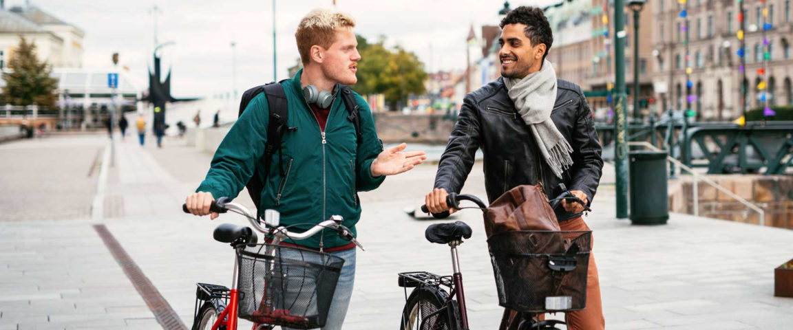 Kaksi nuorta miestä taluttaa polkupyöriä kaupungilla