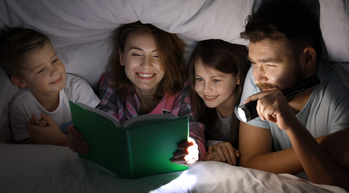 Äiti, isä ja kaksi lasta lukevat peiton alla taskulampun valossa kirjaa.