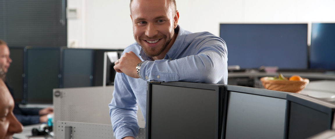 Hymyilevä nuori mies seisoo ja nojaa toimistossa pöytään