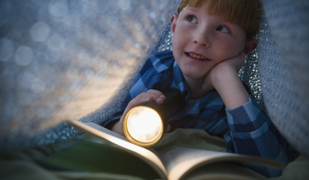 Pieni poika viltistä tehdyssä majassa lukemassa taskulampun valossa