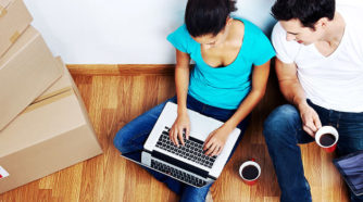 Nuori nainen ja mies istuvat lattialla muuttolaatikoiden vieress sylissään kannettava tietokone
